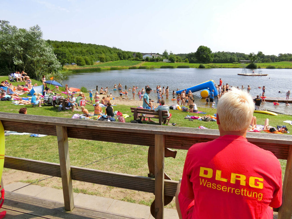 Die DLRG Holmarksee e.V. macht nicht nur beim Kinder-Seefest die Badeaufsicht sondern in der Sommersaison regelmäßig, in den Sommerferien täglich. Auf dem Foto sieht man einen Wachgänger, wie er von der Terrasse herunter blickt auf den See.